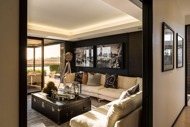 A refined living room with a large cream sectional sofa, dark charcoal feature wall, framed monochrome artwork, and a black trunk-style coffee table. Warm concealed cove lighting outlines the ceiling, while full-height glazing opens toward an outdoor area and brings in daylight. The space has a polished, masculine, contemporary feel with layered neutral furnishings and decorative accents.