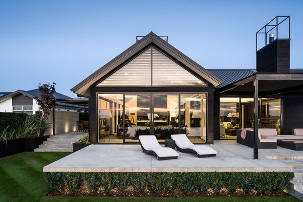 A dusk exterior view of a contemporary single-storey house with a prominent gabled glazed pavilion, dark vertical cladding, and adjoining low-pitched roof volumes. The full-height glazed doors open onto a broad concrete terrace with white outdoor loungers and covered outdoor seating. Landscaped garden beds and neighbouring residential forms frame the property.