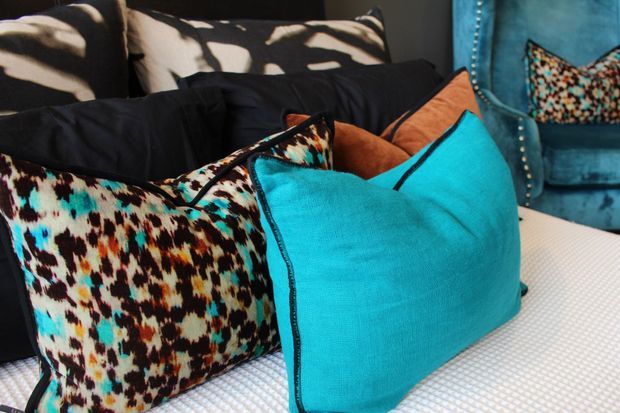 Close-up interior view of a seating area with layered cushions and pillows. Bright turquoise, black, white, orange, and multicoloured patterned textiles create a bold, playful look. A blue upholstered armchair with a patterned pocket or cushion is visible in the background, while the seating surface appears light and textured.