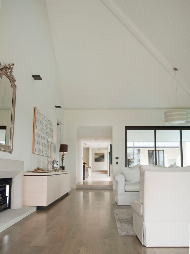 Bright, refined living room with a high sloping white panelled ceiling, pale timber flooring, white upholstered sofas, a minimalist built-in fireplace, and a long low white console. Large black-framed glazing opens the room visually to a light-filled courtyard or adjoining space. A decorative ornate mirror and artwork add subtle traditional accents to the otherwise clean contemporary interior.