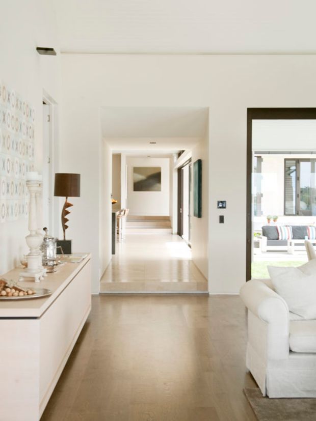 Bright, refined interior view from a living area toward a central hallway and raised adjoining space. The room features white walls and built-in cabinetry, pale polished flooring, dark-framed internal glazing and doors, a sculptural timber table lamp, and a white upholstered sofa in the foreground. A sunlit outdoor or glazed sitting area is visible through the opening on the right.