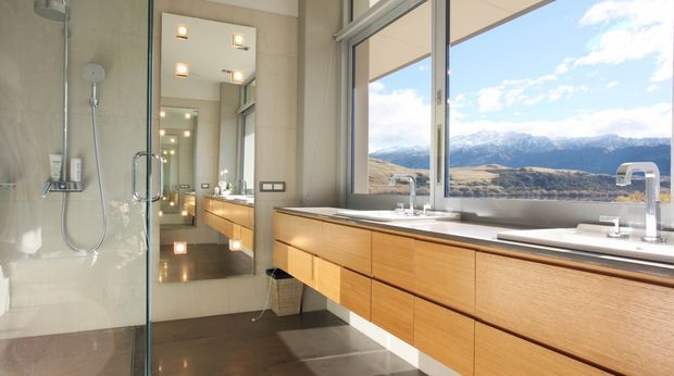 Contemporary bathroom with a long floating timber vanity, pale stone benchtop, large mirror, frameless glass shower, and polished dark floor tiles. Full-height windows provide expansive views of a mountainous landscape, while integrated vanity lighting and chrome tapware create a clean, refined finish.