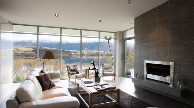 A bright contemporary living room with expansive floor-to-ceiling glazing overlooking a lake and mountainous landscape. The room features a large cream sectional sofa, black-and-white striped lounge chairs, a timber and glass coffee table, recessed ceiling lights, and a sleek dark tiled feature wall with a built-in fireplace and floating hearth.