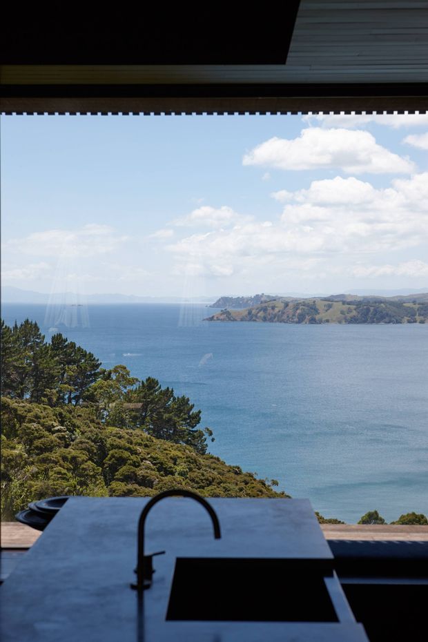 A dark, modern kitchen or outdoor kitchen interior frames a panoramic view across a blue coastal bay and tree-covered headland. In the foreground is a minimalist dark stone or concrete benchtop with an integrated black sink and curved black tap, while a large opening or glazing creates a strong indoor-outdoor connection. The scene has a refined, dramatic coastal atmosphere.