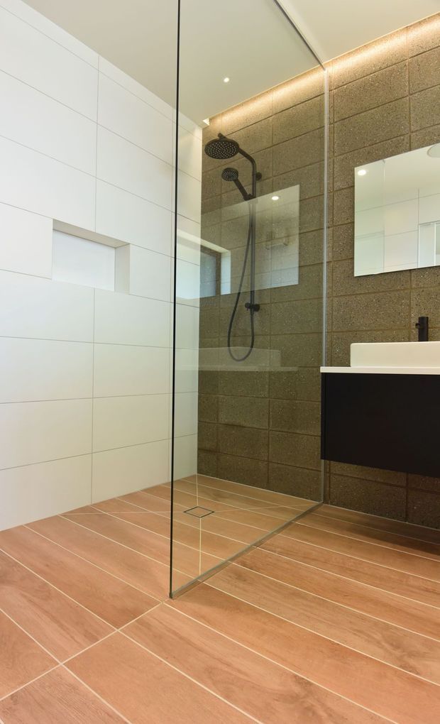 Contemporary bathroom with a walk-in glass shower, large black rain showerhead and handheld fixture, white tiled walls, brown textured masonry-style feature wall, timber-look floor tiles, recessed niche, floating dark vanity and wall mirror. Recessed ceiling lighting and concealed LED strip lighting create a clean, refined finish.