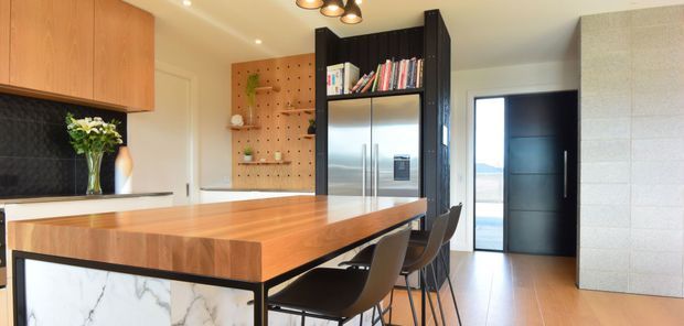 A bright contemporary kitchen featuring a large timber island with waterfall marble-look end panels, dark cabinetry and shelving, stainless-steel refrigerator, black bar stools, and a black textured splashback. A glazed doorway opens toward an exterior view, while a pegboard display wall and exposed blockwork add visual interest.