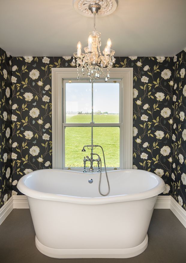 A dramatic traditional bathroom centred on a freestanding white oval bathtub beneath a crystal chandelier. The walls are covered in dark floral wallpaper with white flowers and green foliage, complemented by white decorative trim and a framed sash window overlooking a green field. Chrome bath fittings and a handheld shower are visible.