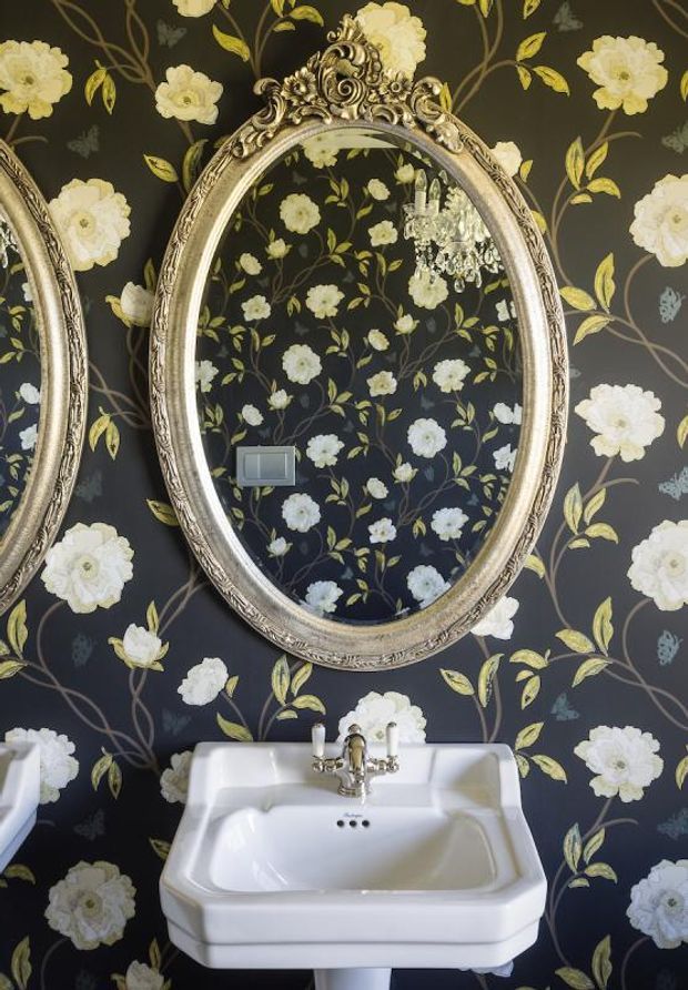 A compact, dramatically styled bathroom featuring a white pedestal basin with traditional chrome taps, ornate silver oval mirrors, and dark floral wallpaper with cream flowers and leafy vines. A crystal chandelier is reflected in the central mirror, adding an opulent vintage character.