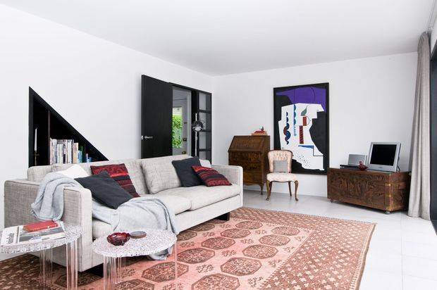 Bright contemporary living room with white walls and floors, a large light-grey sectional sofa, patterned cushions and throw, transparent side tables, and a large muted pink patterned rug. Black built-in triangular shelving and black-framed glazing create contrast, while dark timber antique-style furniture and a colourful abstract artwork add character.
