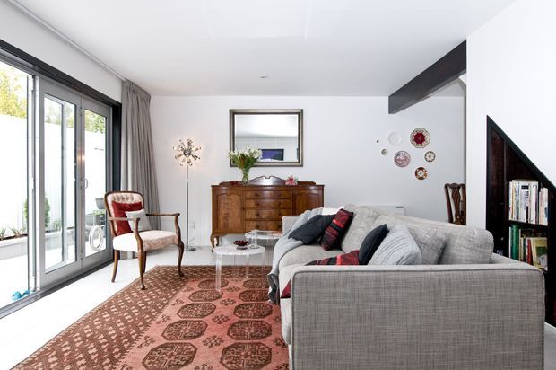 Bright, mid-century-inspired living room with a large grey sectional sofa, patterned area rug, vintage upholstered armchair, timber sideboard, coffee table, and decorative wall accents. Floor-to-ceiling glazed doors open along one side, while a dark exposed beam and built-in shelving add architectural character.