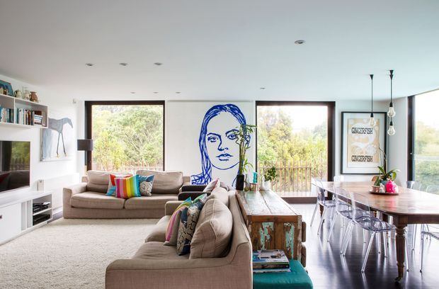 Bright open-plan living and dining area with two large neutral upholstered sofas, a timber dining table with transparent chairs, white built-in shelving and media joinery, and expansive windows overlooking greenery. A bold blue portrait artwork forms a feature wall between the windows, while pendant lights hang above the dining area. The room has a relaxed contemporary feel with colourful cushions and a mix of timber and painted finishes.