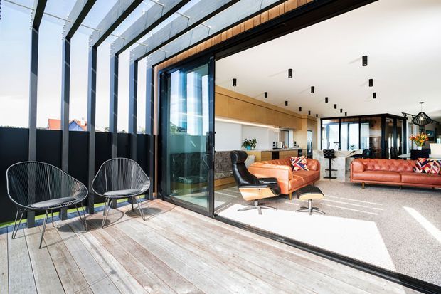 Bright contemporary open-plan living area extending onto a timber deck through large sliding glass doors. The interior features a white ceiling with repeated recessed black downlights, warm timber cabinetry and wall finishes, a kitchen, orange-red sofas, and a black leather lounge chair. The deck has a dark metal-framed glazed roof or pergola structure and outdoor wire-frame seating.