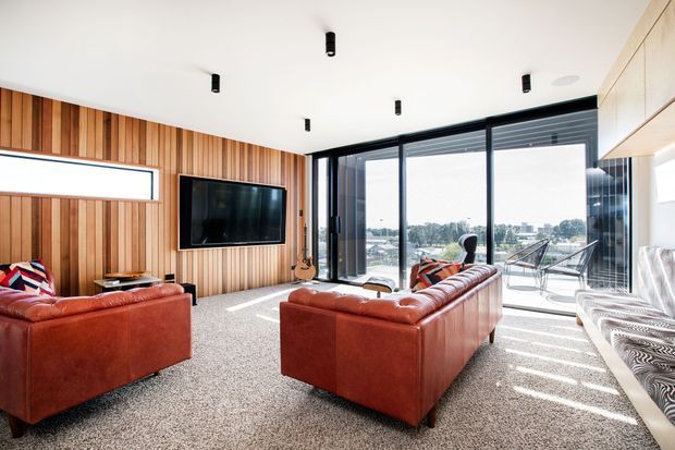 Bright contemporary living room with rich timber wall panelling, red-brown leather sofas, a wall-mounted television, and full-height black-framed glazing opening to an outdoor terrace. A pale built-in cabinetry wall lines the right side, while strong daylight creates patterned shadows across the carpeted floor.