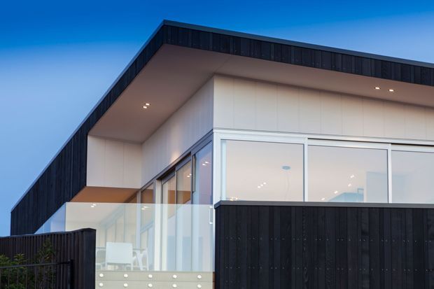 Exterior architectural photograph of a contemporary multi-level home at dusk, featuring crisp geometric forms, black vertical cladding, large white-framed glazed openings, a broad roof overhang, and a glass-railed balcony or terrace.