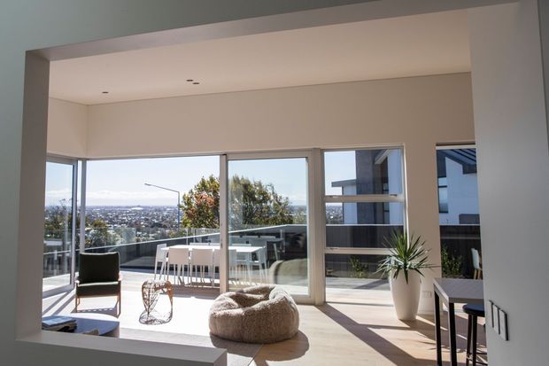 Bright contemporary living room framed by a large interior opening, with floor-to-ceiling glazing opening onto a balcony or terrace and views over a suburban landscape. The room includes a low sofa, sculptural lounge chair, beanbag, small side table, indoor plant, and a compact dining area. Pale timber flooring, white walls, slim dark window frames, and abundant natural light create an airy, minimal interior.