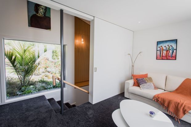A bright contemporary living room overlooking a planted outdoor area through large glazing. The room features dark charcoal carpet, white walls, a cream sectional sofa with orange and patterned throws, sculptural white nesting coffee tables, and a stairwell screened by a slim dark-framed glass balustrade. A warm timber-lined wall with pendant lighting adds contrast, while colourful artwork and a tall floor lamp provide decorative accents.