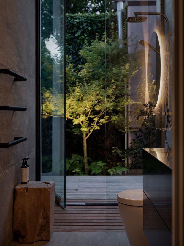 Moody, luxurious bathroom interior with a frameless glass shower enclosure opening toward a timber deck and lush planted garden. The room features dark textured wall finishes, a sculptural timber stool, black minimalist fixtures and shelving, a freestanding toilet, dark vanity cabinetry, and warm integrated lighting. Reflections in the glazing add depth and connect the bathroom visually with the greenery outside.