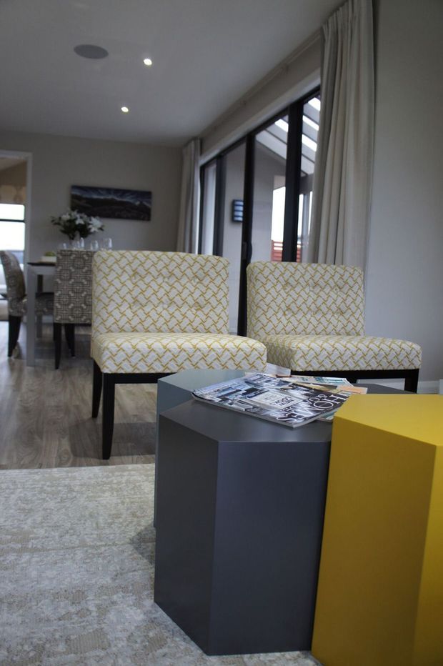 A contemporary living room with two patterned upholstered lounge chairs positioned beside dark geometric side tables and a bright yellow cube table. A pale area rug, timber-look flooring, neutral walls, recessed ceiling lights, artwork, and full-height black-framed glazed doors with curtains complete the room. A dining area is visible in the background.