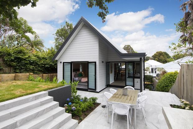 Exterior view of a compact contemporary gable-roof home with white horizontal weatherboard cladding, dark window and door frames, and a paved outdoor dining terrace. Broad concrete steps lead up to the landscaped patio, while glazed openings reveal a small interior living area. The setting includes established suburban planting and neighbouring homes.