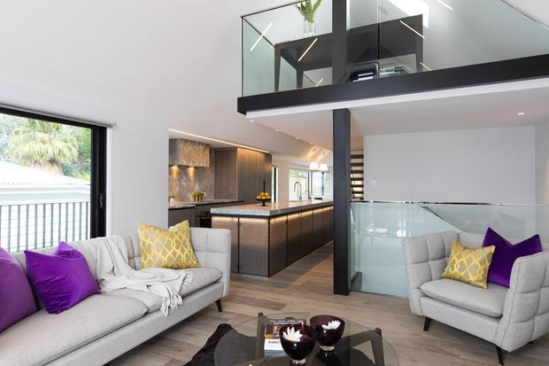 Bright contemporary open-plan living space with two light upholstered sofas, colourful purple and yellow cushions, a black glass coffee table, and a dramatic double-height void. A modern kitchen with dark timber cabinetry and a textured island sits behind the living area, while a glass balustrade and mezzanine overlook the room.
