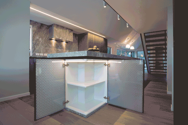 A contemporary open-plan kitchen with a large island featuring patterned panelled cabinetry and an open corner display niche. The island has a pale stone benchtop, dark timber cabinetry, a textured stone-look splashback, integrated under-bench lighting, and a small fruit bowl. Warm linear and track lighting illuminate the kitchen, while a dark timber open-riser stair or slatted partition is visible to the right. Dark timber flooring and neutral finishes create a refined, modern interior.