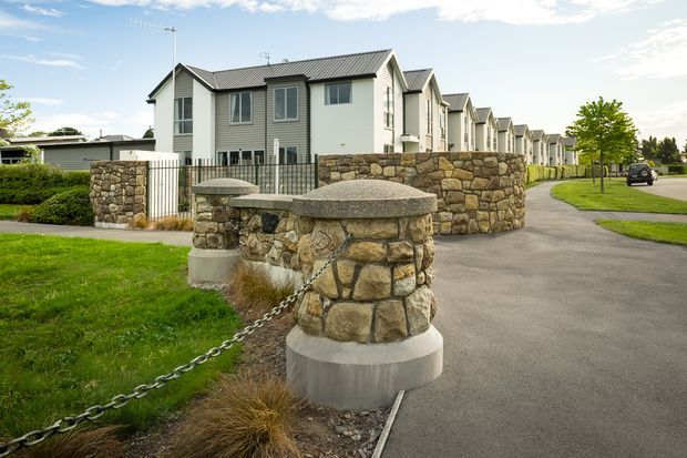 Exterior view of a contemporary multi-unit residential development with repeated gabled townhouse forms, pale rendered walls, dark pitched roofs, and prominent rough natural-stone boundary walls and chain-linked stone bollards. The landscaped verge, paved internal driveway, and uniform row of homes are clearly visible.