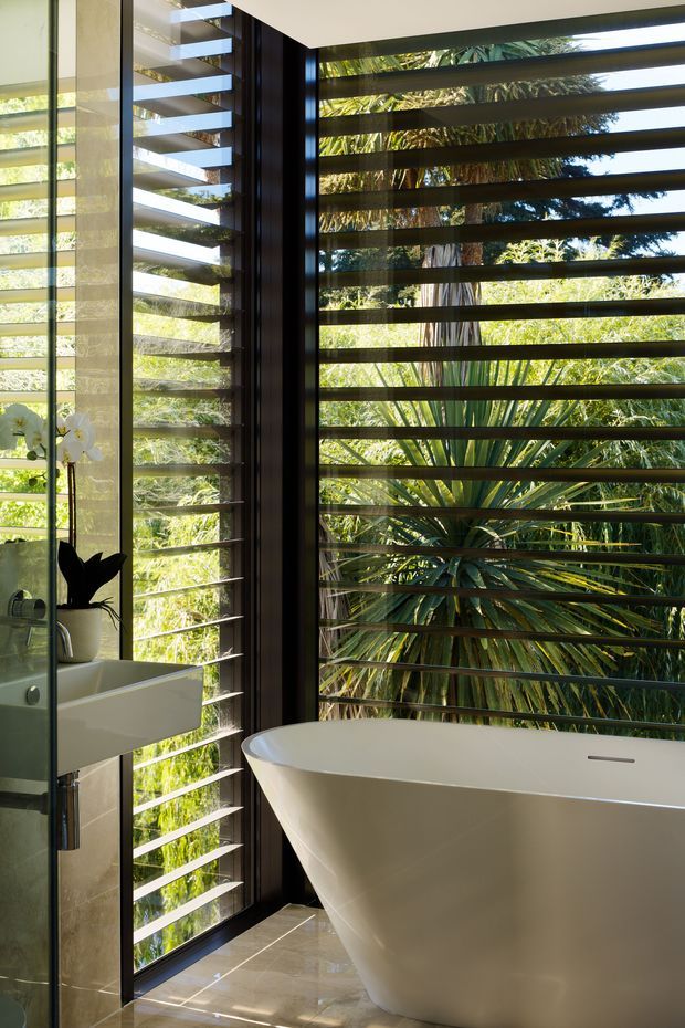 A bright, refined bathroom with a freestanding white bathtub and wall-mounted rectangular basin set against full-height dark-framed glazing. Horizontal external louvres provide privacy and sun control while allowing views to dense tropical greenery. Pale stone or porcelain floor and wall surfaces, dark window frames, and abundant filtered daylight create a serene indoor-outdoor atmosphere.