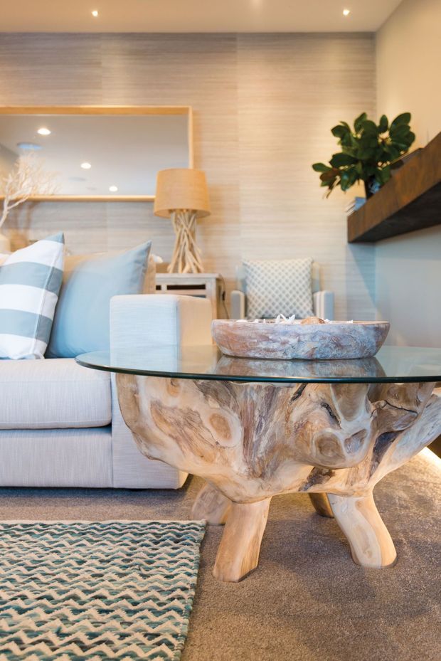 A warmly lit contemporary living room featuring a pale upholstered sofa with blue and white striped cushions, a sculptural natural timber coffee table with a glass top, a textured area rug, a woven accent chair, and a timber-framed wall mirror. A floating dark timber shelf with a potted plant adds contrast against the light horizontal wall finish.