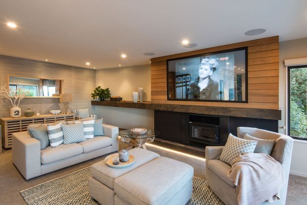 A refined living room centered on a large built-in media wall with a wall-mounted television, dark fireplace surround, and horizontal timber panelling. The room includes a pale upholstered sofa, armchair, oversized ottoman, patterned rug, glass coffee table, timber slatted console, and recessed ceiling lighting. Large windows bring in garden views and natural light.