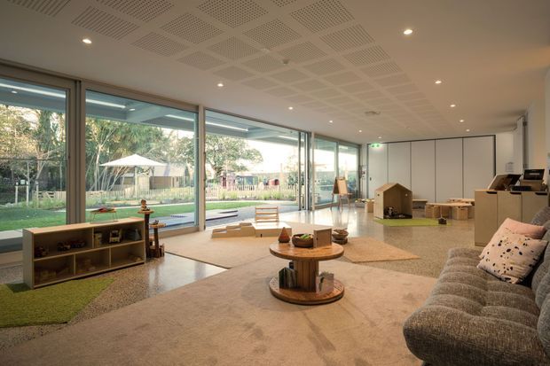 A bright, spacious contemporary living room with expansive floor-to-ceiling glazing overlooking a landscaped outdoor area. The room features a large textured sectional sofa, layered neutral rugs, low timber coffee tables, open shelving, children's play furniture, and a patterned acoustic ceiling. Sliding glass doors create a strong connection to the garden and outdoor play space.