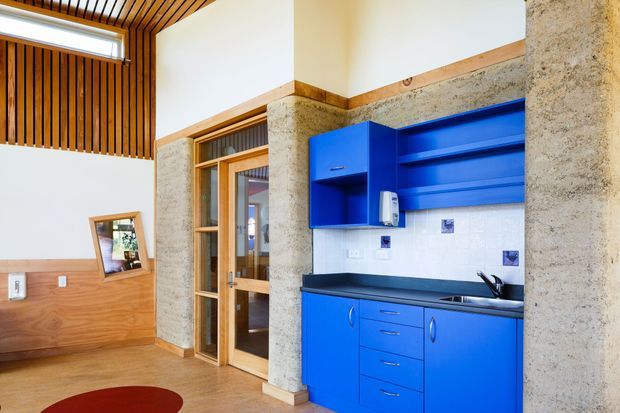Bright compact kitchenette integrated into an open interior, with vivid blue fixed cabinetry, dark benchtop, white tiled splashback, timber-framed glazed door, exposed rough concrete walls, and a warm timber slatted ceiling and wall panel. The space has a distinctive utilitarian yet colourful contemporary character.