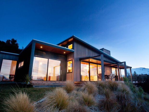 Exterior dusk view of a contemporary single-storey house on a landscaped, sloping site. The building has dark vertical cladding, extensive floor-to-ceiling glazing, projecting roof forms, covered outdoor living areas, and warm interior lighting. Native tussock-style planting surrounds the house, with distant mountainous terrain visible beyond.