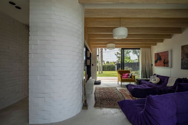 A contemporary living room with a large curved white-painted brick feature wall, exposed timber ceiling beams, polished concrete flooring, and expansive glazed doors opening to a garden. Deep purple sectional seating, a mustard armchair, patterned rug, and artwork create a bold, eclectic interior. A fireplace or built-in joinery is partially visible beside the curved wall.