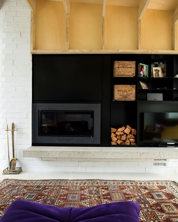 Interior view of a living room feature wall with a black built-in media and fireplace unit set into white-painted brickwork. The joinery includes open shelving, a television, decorative objects and stacked firewood, with pale timber display cupboards above and a concrete hearth ledge below. A richly patterned area rug and the edge of a deep purple sofa occupy the foreground.