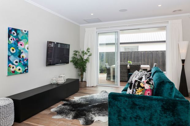 A bright contemporary living room with a deep teal velvet sofa, black low-profile media console, wall-mounted television, colourful abstract artwork, indoor plants, patterned cushions, and a black-and-white hide rug. Full-height glazed doors open to a small paved courtyard, while white curtains, recessed ceiling lights, and pale neutral walls create an airy, polished interior.