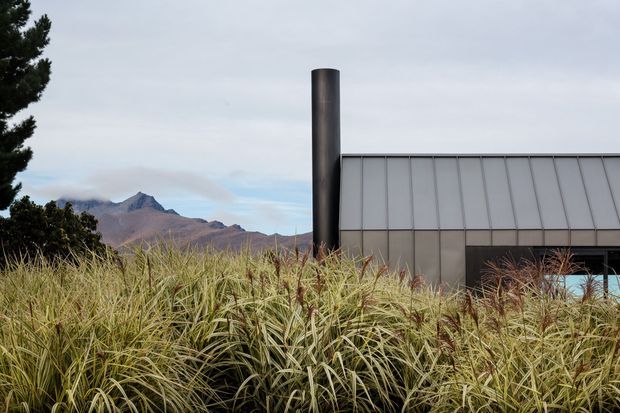 Exterior view of a contemporary building partially screened by tall ornamental grasses. The structure has a simple rectangular form, a large dark cylindrical chimney or flue, and a grey vertically articulated metal-clad facade. Rugged mountains are visible in the background beneath an overcast sky.