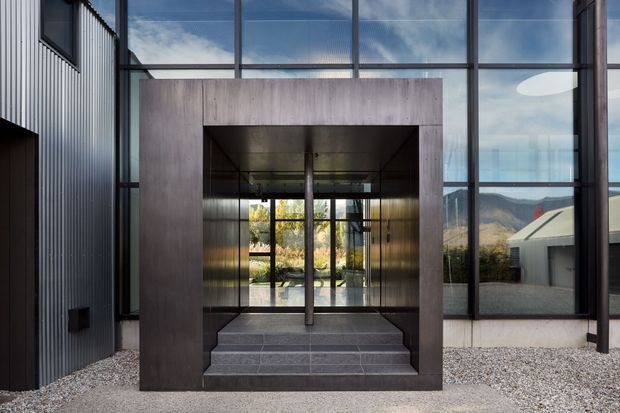 A contemporary building entrance viewed from outside, featuring a dramatic deep rectangular portal clad in dark timber or wood-look panels, broad charcoal tiled steps, and a fully glazed double-height facade. Vertical dark corrugated cladding frames the left side, while the glazing reflects sky and reveals landscaped interior space and distant mountains beyond. The entry is set against a pale gravel forecourt and has a strong minimalist, geometric composition.
