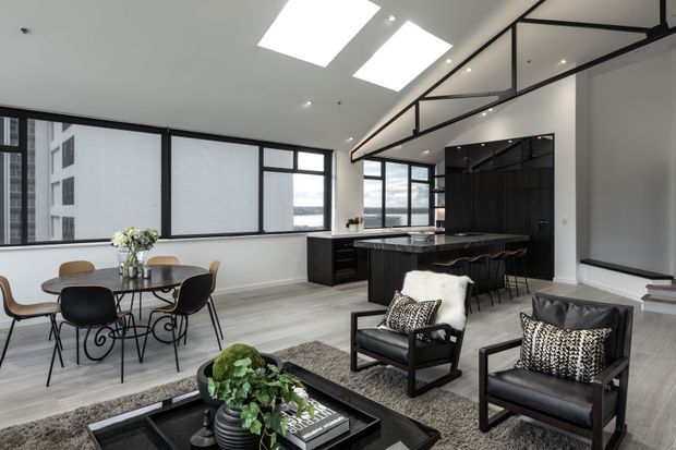 Open-plan contemporary apartment interior combining a dining area, kitchen and living space. The room features extensive black-framed glazing, a high ceiling with skylights, pale timber flooring, a dark kitchen island and cabinetry, and black leather lounge chairs around a low coffee table. A glass-and-black-metal mezzanine or balustrade detail runs along the upper wall.