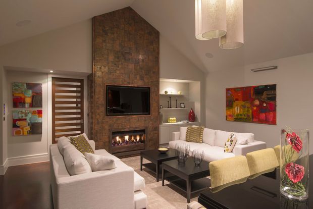 A refined living room with a vaulted ceiling and a tall rustic stone-clad fireplace wall incorporating a television and linear gas fireplace. Two light sofas, pale upholstered dining or occasional chairs, a dark coffee table, built-in shelving, colourful artwork, and warm pendant lighting create a comfortable contemporary setting.