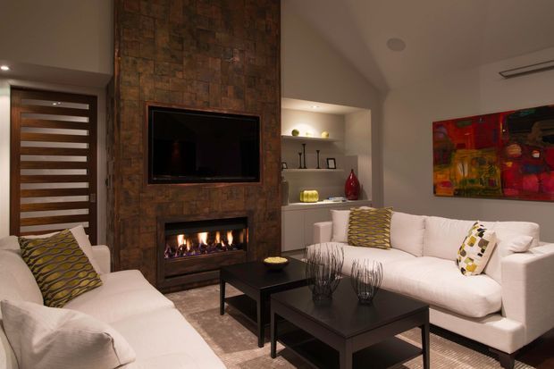 A warm, contemporary living room with two large white upholstered sofas arranged around black rectangular coffee tables. A floor-to-ceiling stacked stone feature wall incorporates a wall-mounted television and built-in gas fireplace, with recessed display shelving and cabinetry beside it. A timber-and-glass front door, colourful abstract artwork, patterned cushions, and soft recessed lighting add visual interest.