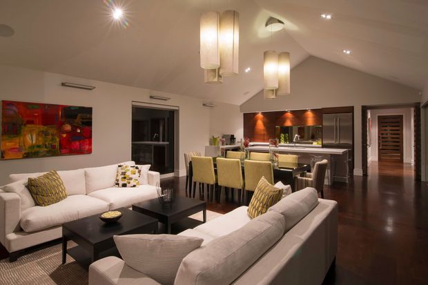 Open-plan contemporary living and dining area with a modern kitchen in the background. The room features cream upholstered sofas, a black coffee table, a dark timber floor, a dining table with upholstered chairs, dark kitchen cabinetry, polished benchtops, recessed lighting, and prominent pendant lights. A colourful abstract artwork adds a vivid accent.