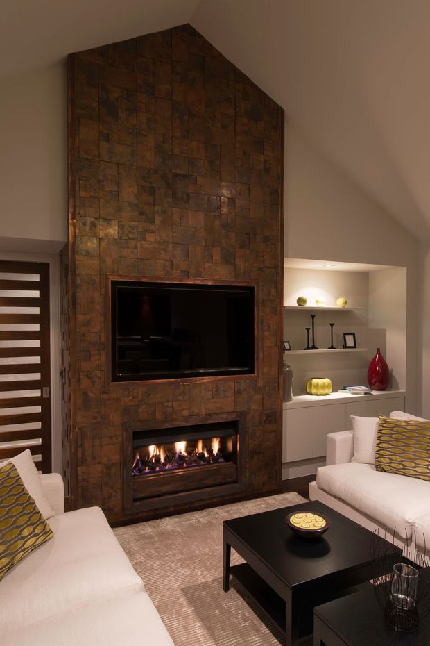 A warm contemporary living room centred on a tall, pitched-top feature wall clad in dark copper-toned textured panels. The wall incorporates a wall-mounted television above a built-in linear fireplace. White sofas with patterned cushions face a black rectangular coffee table, while recessed shelving displays small decorative objects and framed art. A modern slatted timber-and-glass entry door is visible at left.