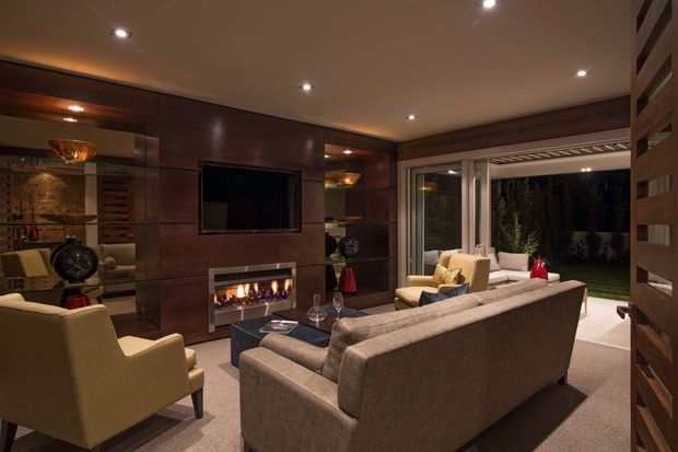 A sophisticated, warmly lit living room with dark timber wall panelling, a wall-mounted television, linear fireplace, plush seating, and full-height glazed doors opening to a garden. Recessed ceiling lighting, built-in shelving, and neutral upholstery create a refined contemporary entertaining space.