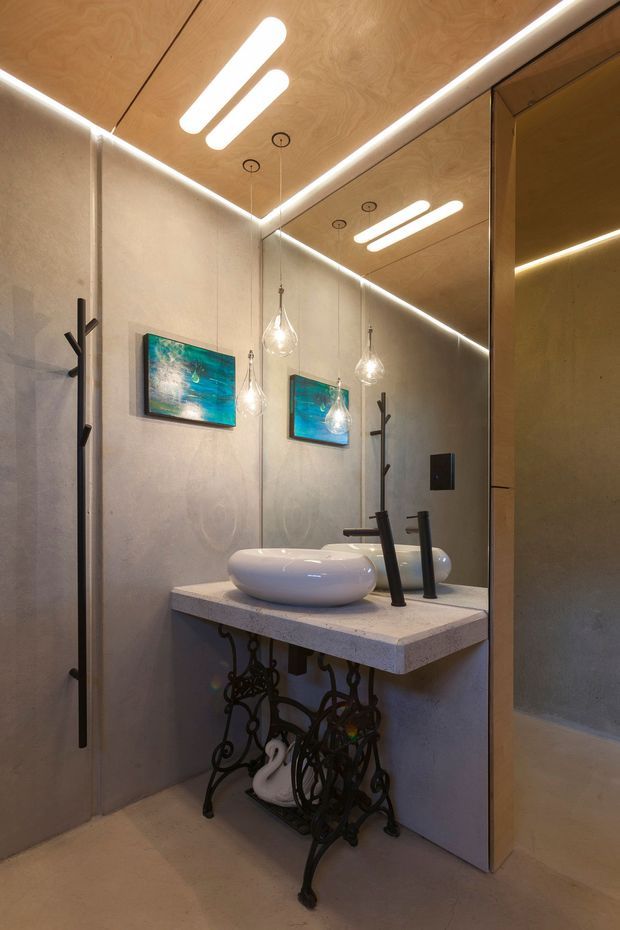 A compact, moody bathroom with a floating-style concrete or stone vanity slab, two white vessel basins, matte black tapware, and ornate black wrought-iron supports beneath. The walls and floor have a warm grey plaster or concrete finish, while a timber-lined ceiling incorporates recessed linear lighting. Large mirrors reflect the vanity and pendant lights, with turquoise artwork adding a small colour accent.