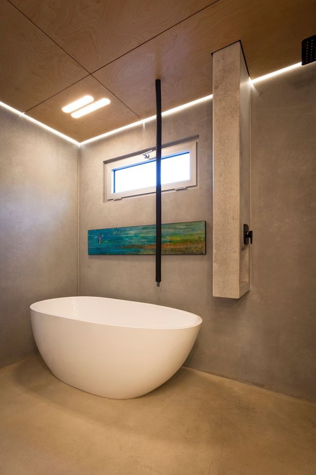Compact contemporary bathroom featuring a freestanding white oval bathtub, seamless warm grey concrete-like walls and floor, a plywood ceiling with linear recessed lighting, a narrow horizontal window, and minimalist black hardware. A slim vertical shower fixture and a colorful horizontal artwork add contrast.