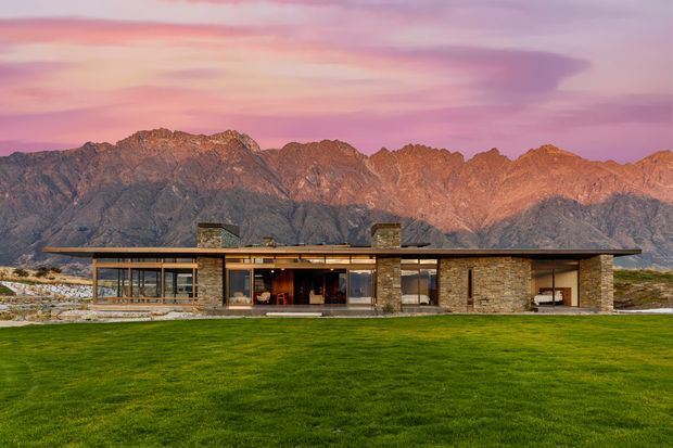 A low, single-storey contemporary residence set against a rugged mountain range. The home has a broad flat roof with deep overhangs, extensive floor-to-ceiling glazing, timber-framed window walls, and substantial stacked-stone wall elements and chimneys. A large green lawn occupies the foreground, while the mountains and pink sunset sky form the background.