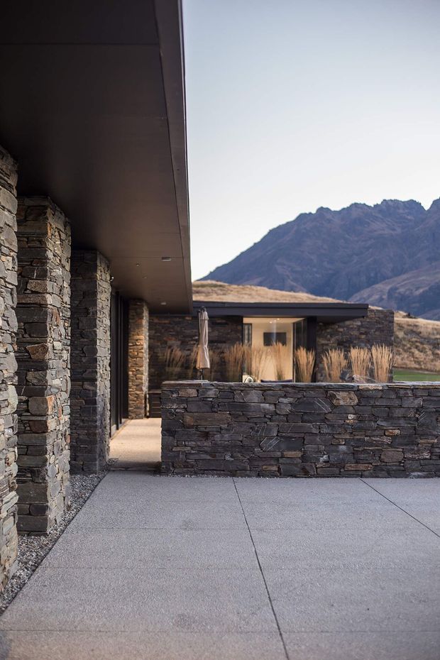 Exterior view of a low, contemporary mountain residence with extensive dark stacked-stone walls, deep roof overhangs, and flat-roofed forms. A concrete-paved terrace and stone privacy wall lead toward a glazed courtyard or entry area, with dry ornamental grasses and rugged mountains visible beyond.