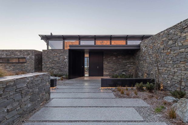 A low-slung modern residential entrance composed of dark stacked-stone walls, a recessed black entry door, and a slim glazed clerestory beneath a projecting roof canopy. Broad concrete stepping slabs lead through a landscaped courtyard with raised stone planters and sparse ornamental grasses.