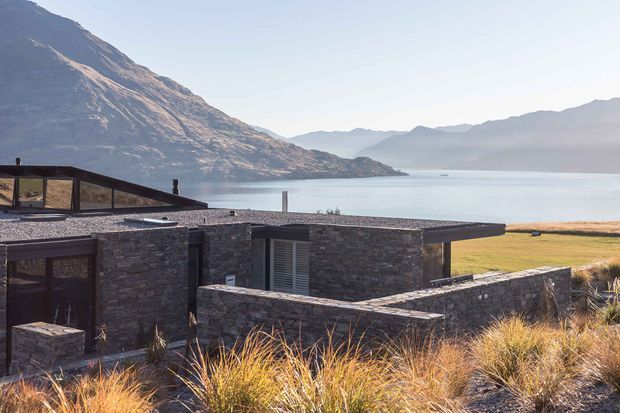 A low-slung contemporary stone-clad residence set against a dramatic alpine lake and mountain landscape. The building has a flat roof, dark horizontal glazing, exposed stone walls, and landscaped grasses around the terrace and entry areas.