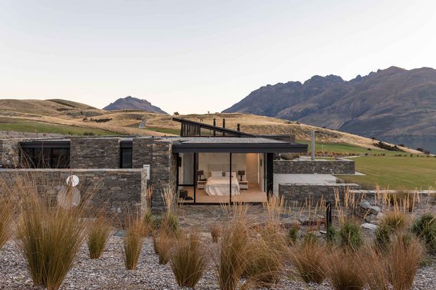 A low-profile contemporary house set within a dry, mountainous landscape. The dark stone-clad facade features broad glazing, a covered outdoor terrace, and a flat or very low-pitched roofline. Ornamental grasses and gravel landscaping surround the building, while a bedroom is visible through the central glazed opening.